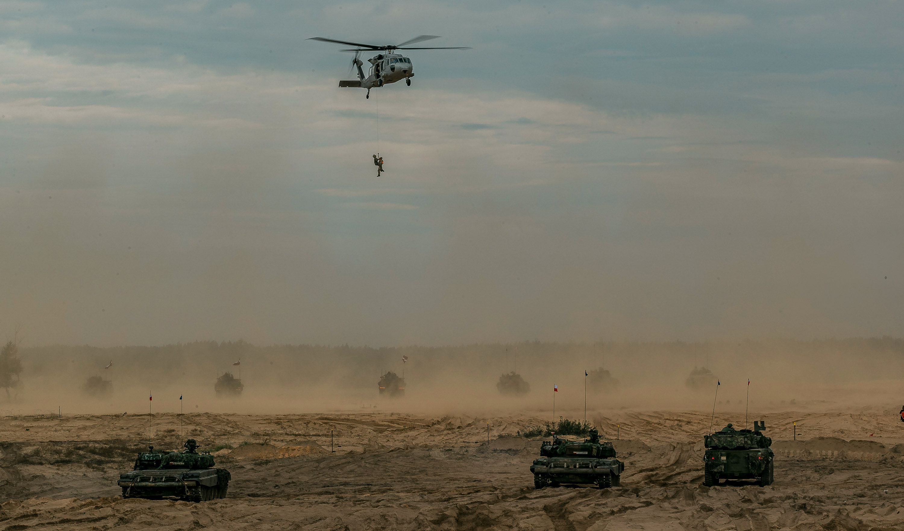 a helicopter lowers a soldier over a dusty training ground filled with military vehicles