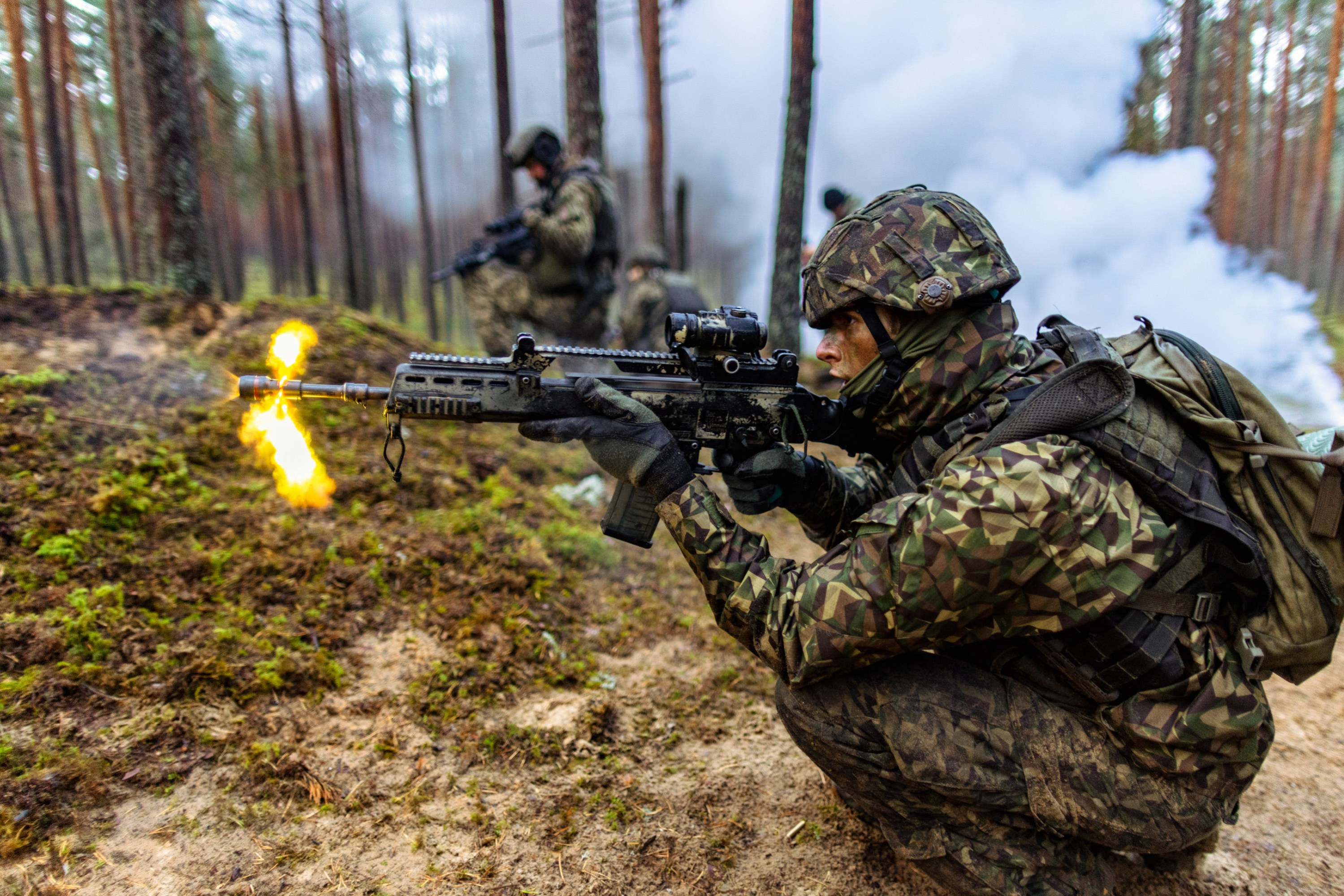 soldier in full camoflage firing a gun in a wooded area with smoke and several other soldiers out of focus behind him